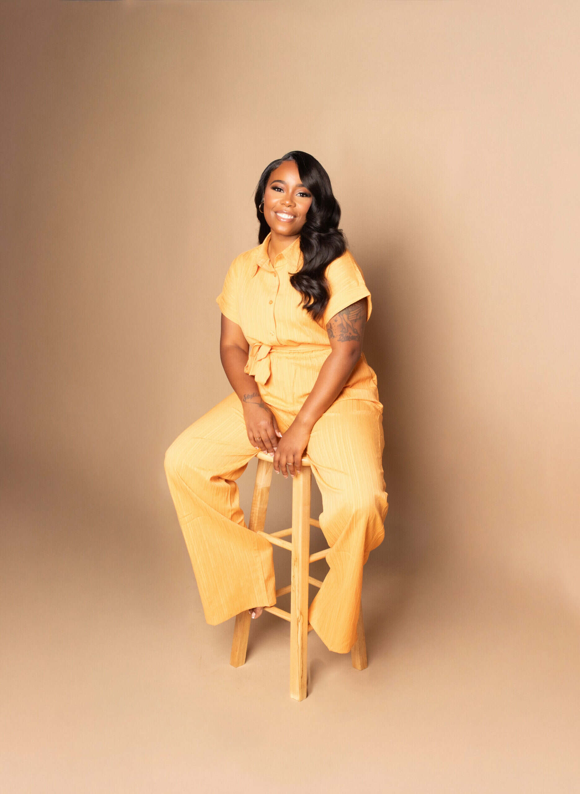 A woman sitting on a wooden stool wearing a yellow outfit against a beige background.