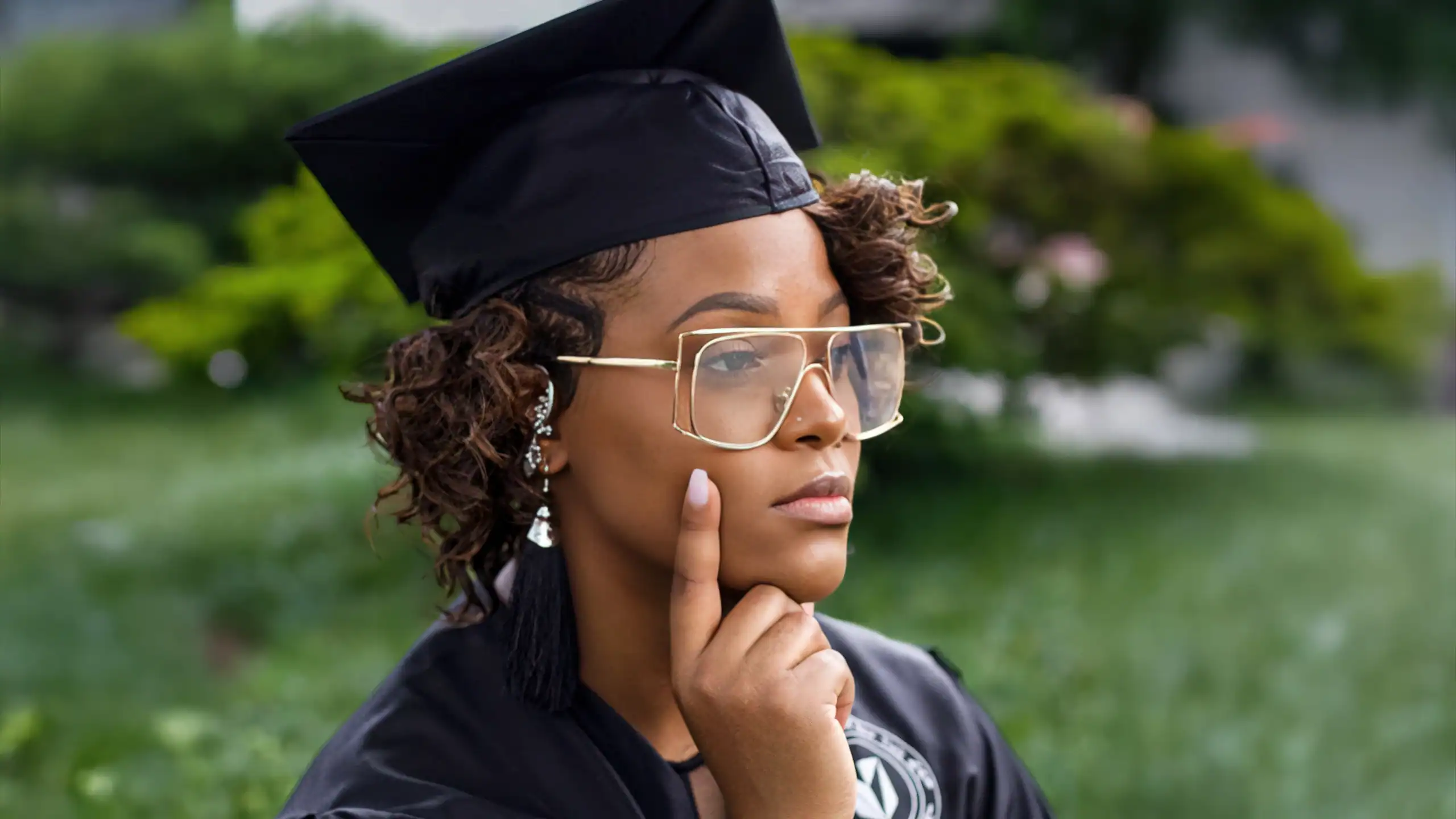 Elegant young woman in graduation cap and gown, outdoors, thoughtful pose, celebrating academic achi.