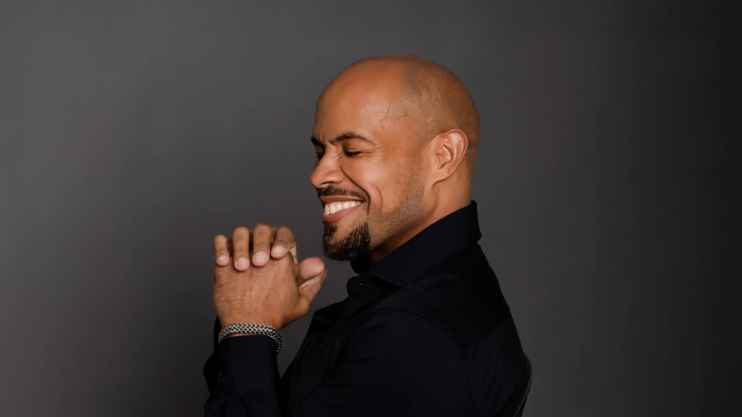 A smiling man with a beard and closed eyes, wearing a black shirt, against a dark gray background.