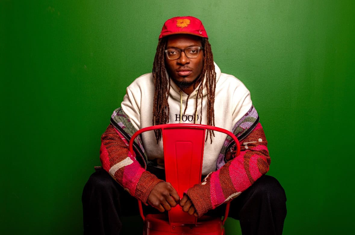 Portrait of a man with dreadlocks wearing glasses and a red cap, sitting on a red chair against a gr.