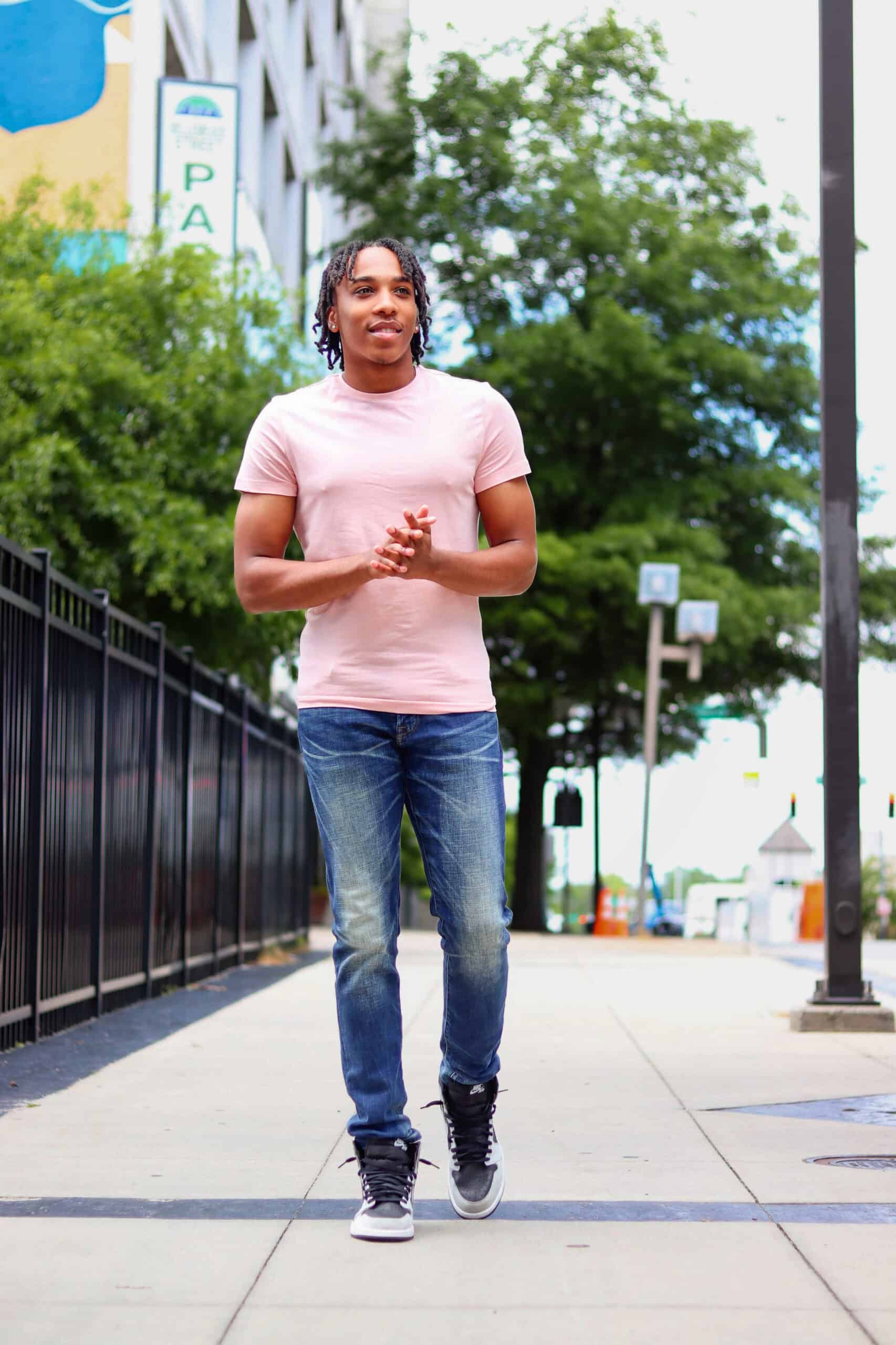 Young man walking confidently on city sidewalk with trees and buildings in background.