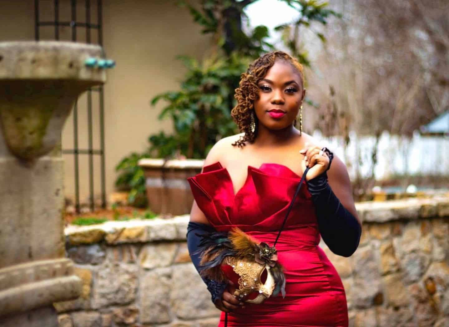 Elegant woman in a red evening gown with black gloves, standing outdoors with a stone wall and green.