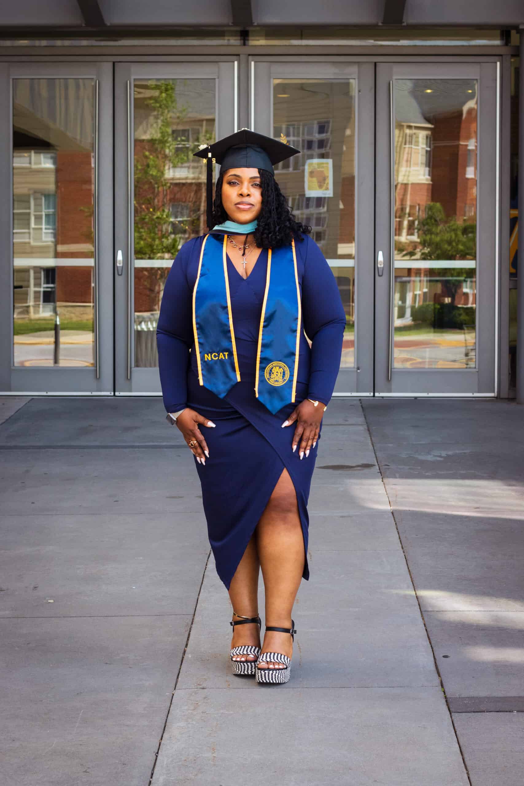 Woman in graduation cap and gown standing outdoors, celebrating academic achievement.