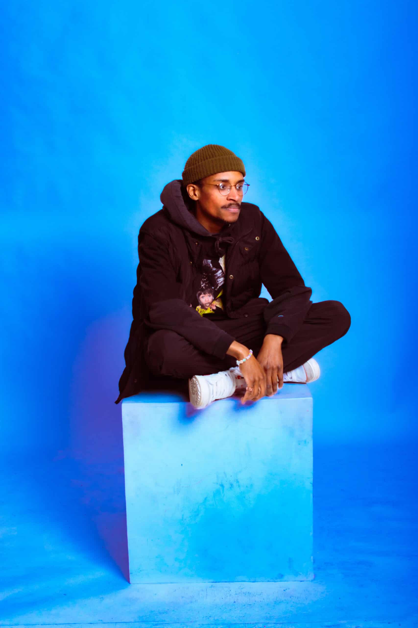 Modern portrait of a man sitting on a blue block with a colorful background.