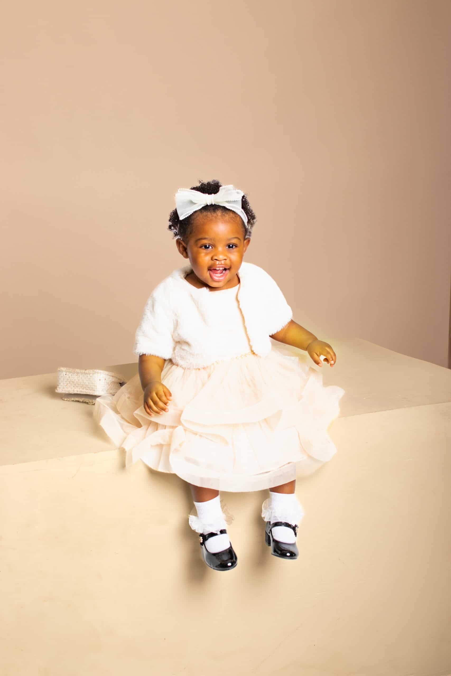 Baby girl in white dress with bow, sitting and smiling, showcasing adorable fashion.