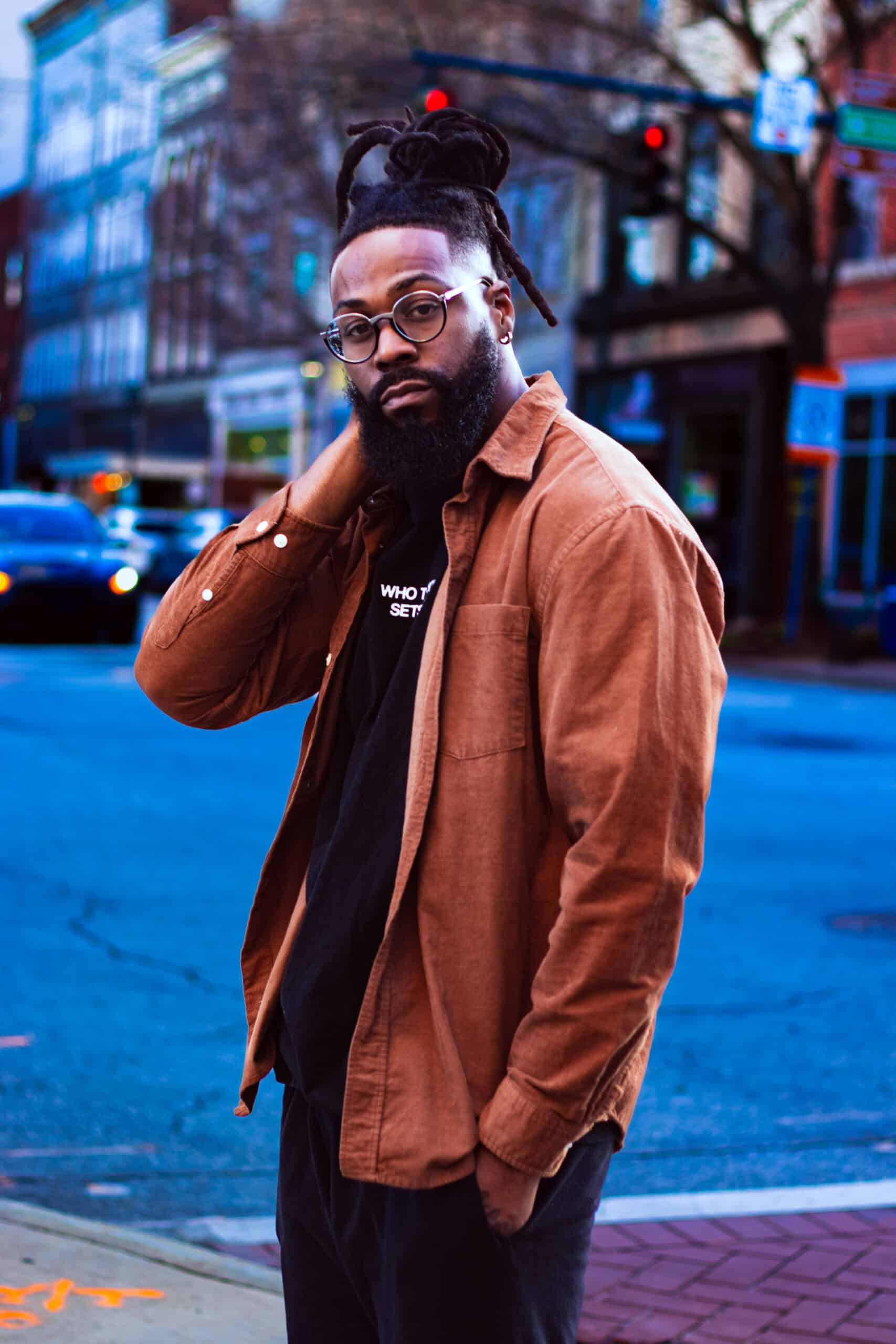 Young man with glasses and dreadlocks standing on city street, urban fashion style.