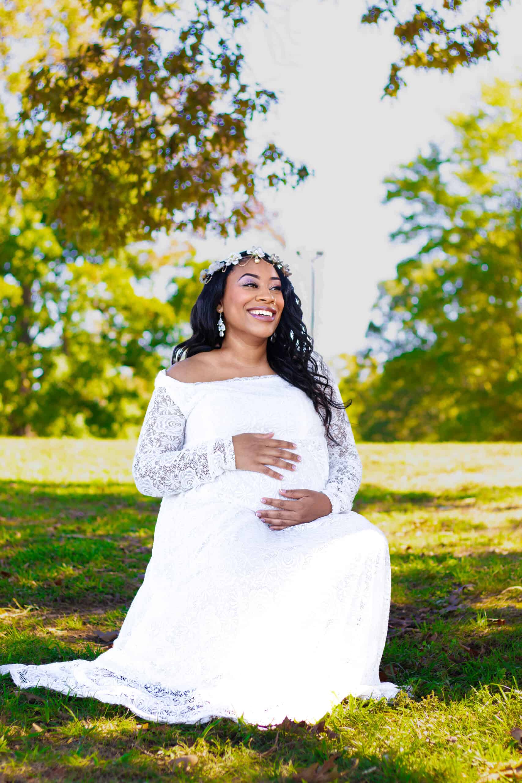 Pregnant woman in a white lace dress sitting on grass in a park, smiling and holding her belly. Beau.