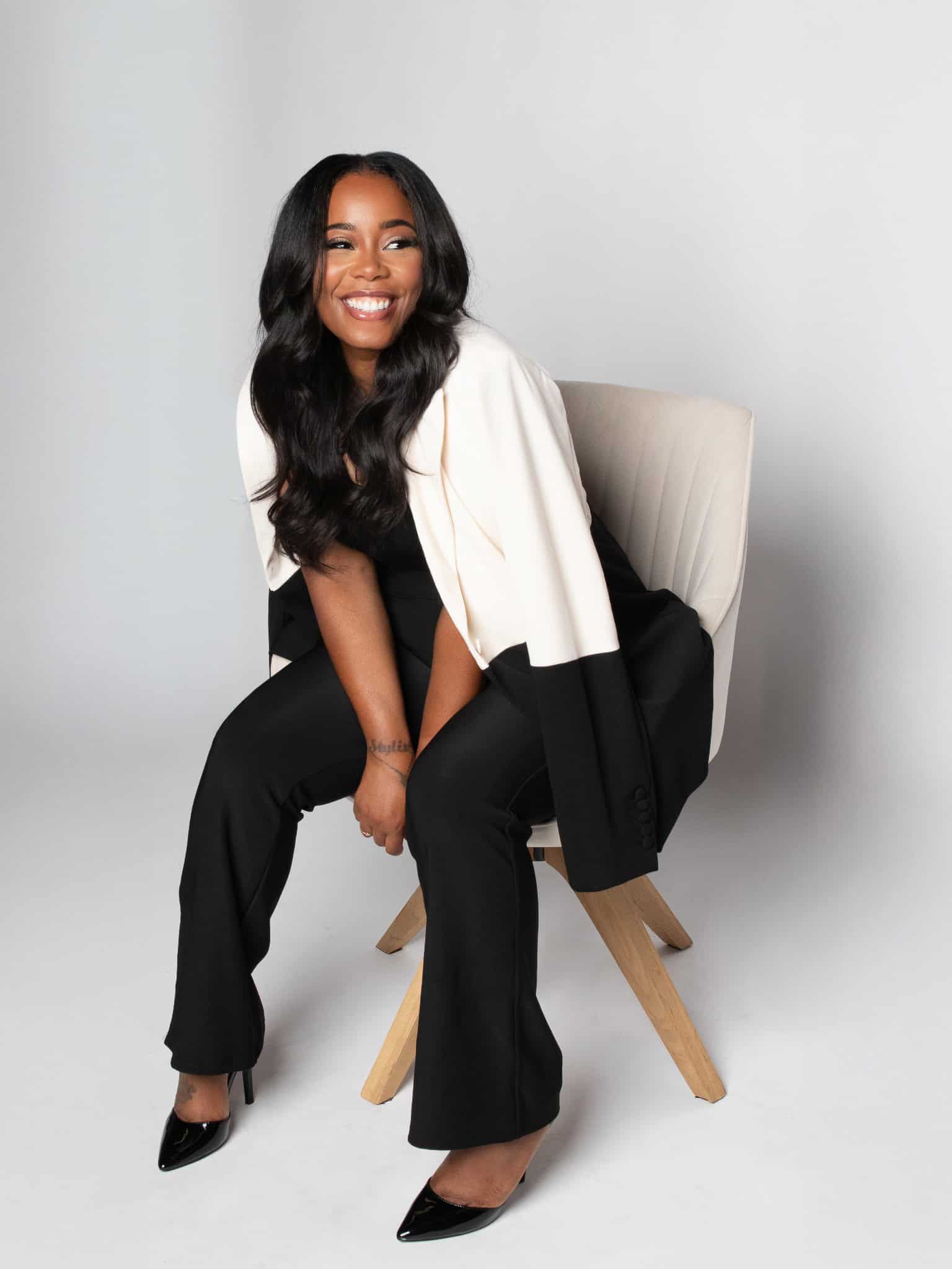Confident woman in black and white outfit sitting on a modern chair, smiling at the camera.