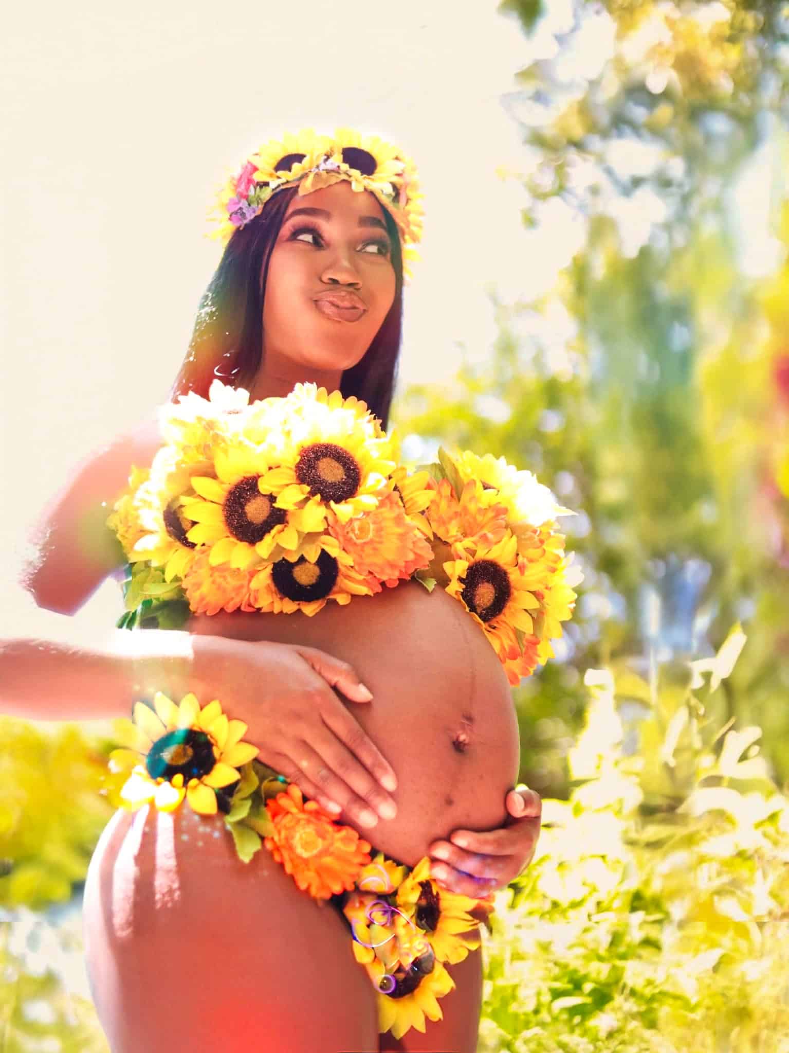Woman with sunflower crown and flowers on belly, outdoor sunny setting.