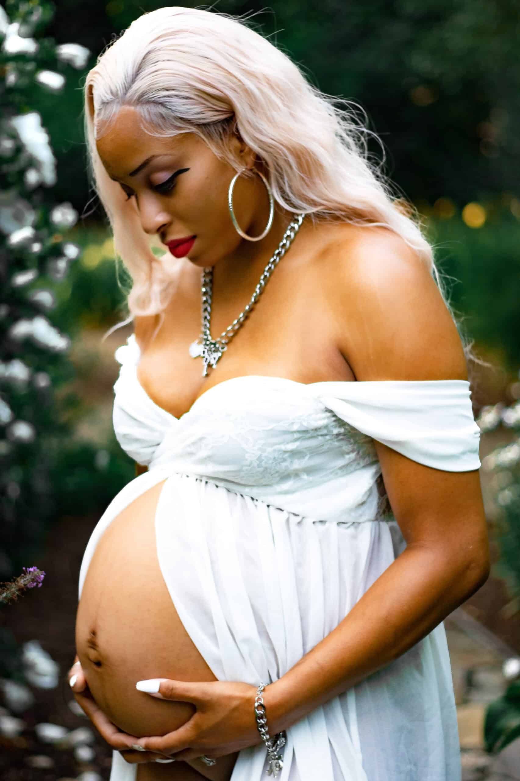 Pregnant woman with blonde hair in a white dress holding her belly outdoors.