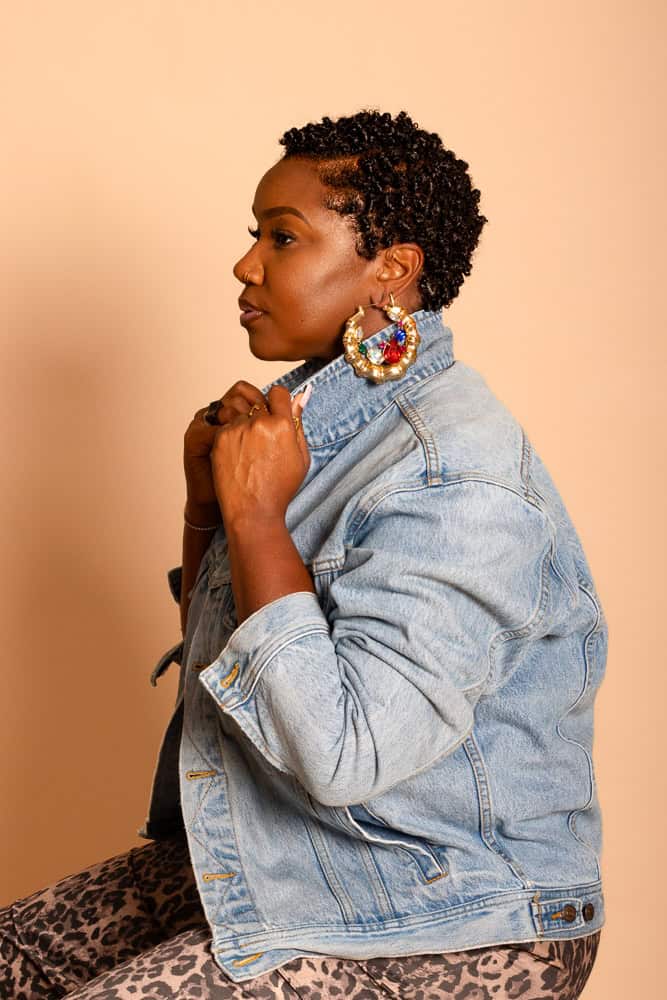 Woman with natural curly hair, denim jacket, and vibrant earrings posing confidently.