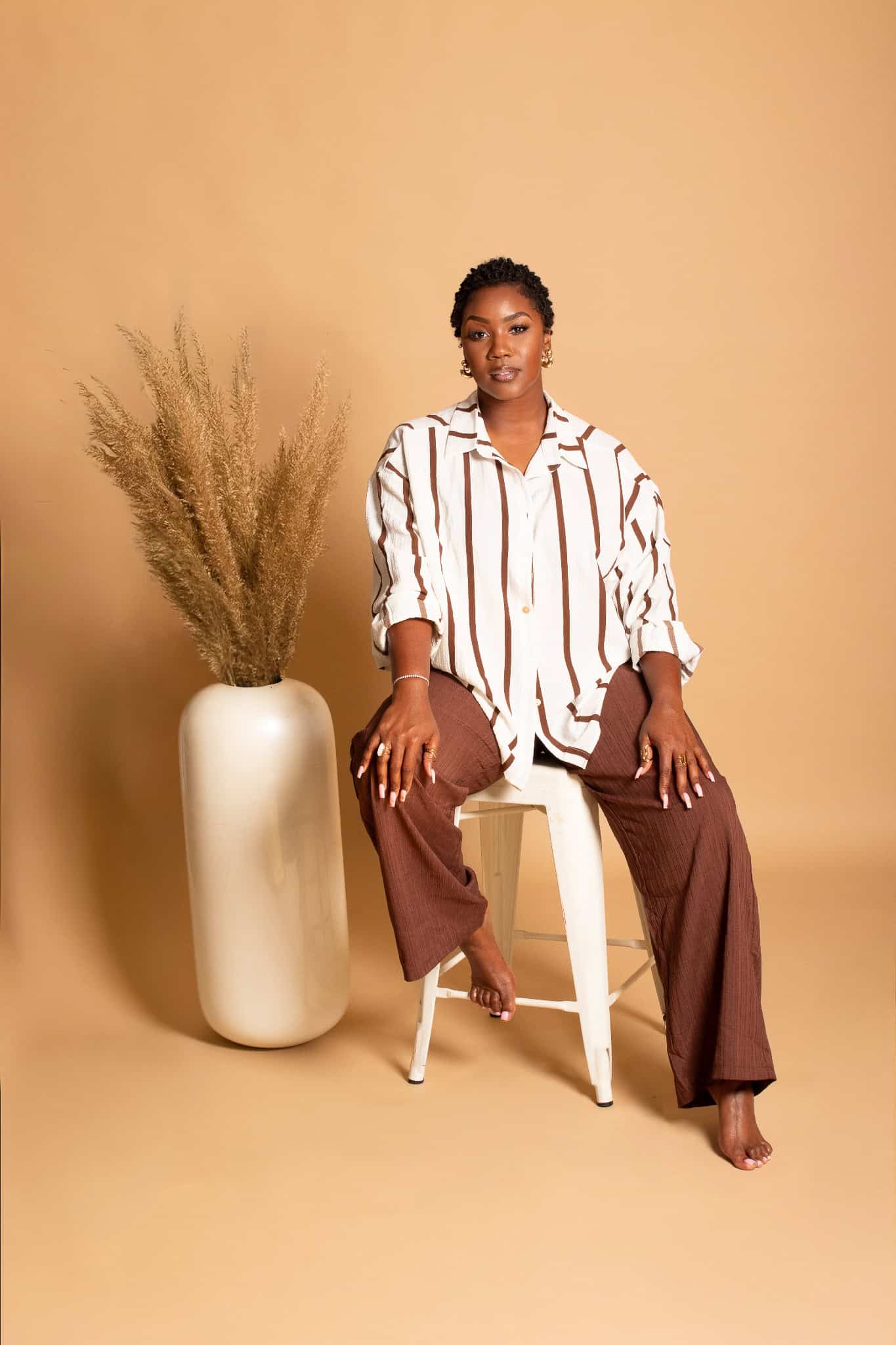Confident woman seated on a chair with a vase of dried plants against a beige background.