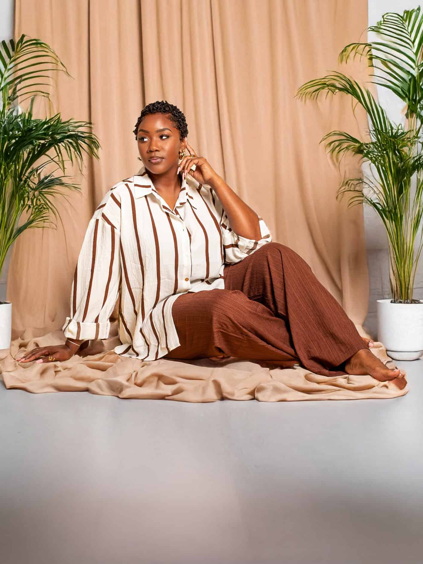 Elegant woman seated on the floor with lush green plants and beige curtains in the background.