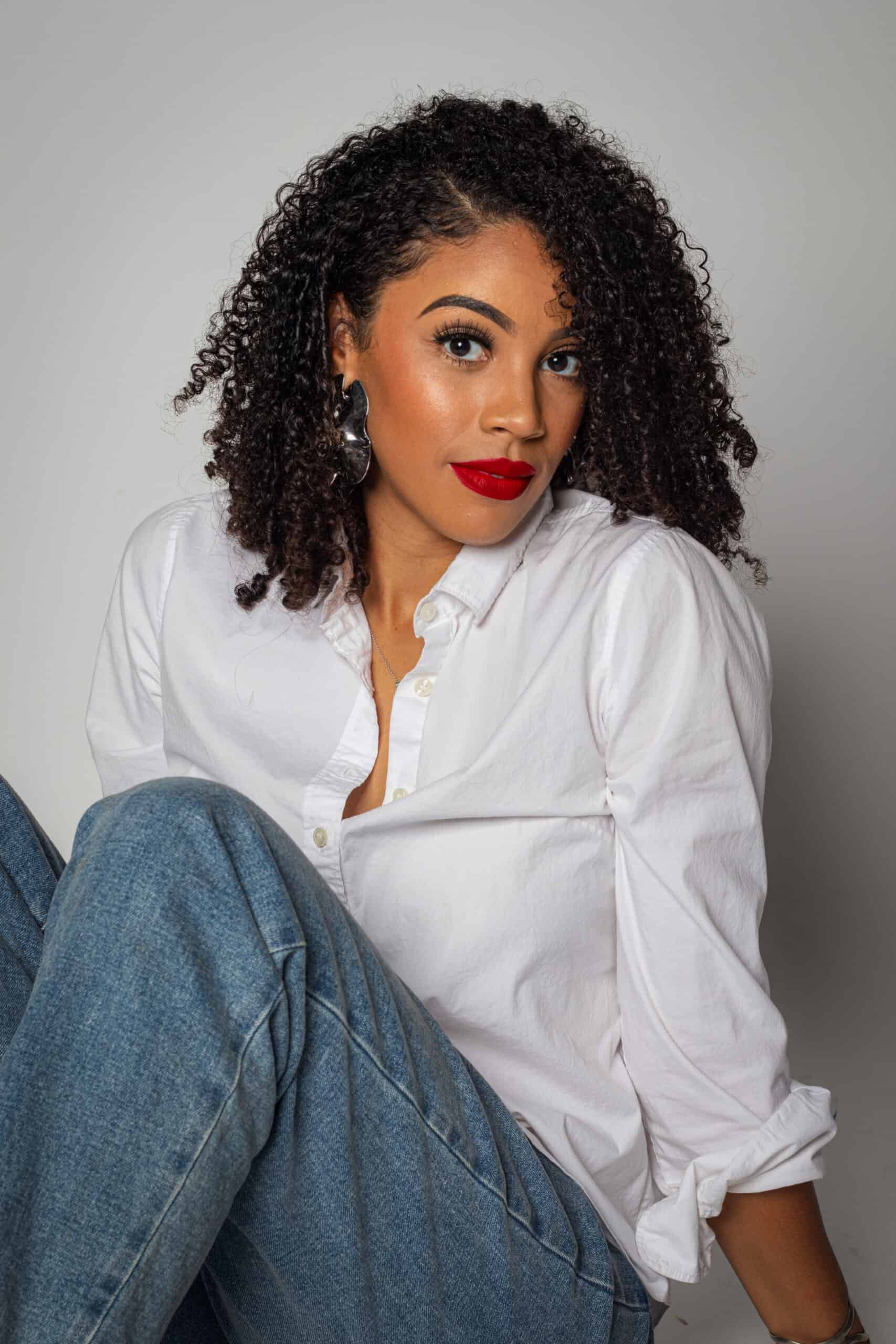 Elegant woman with curly hair wearing a white shirt and jeans, posing confidently against a neutral.