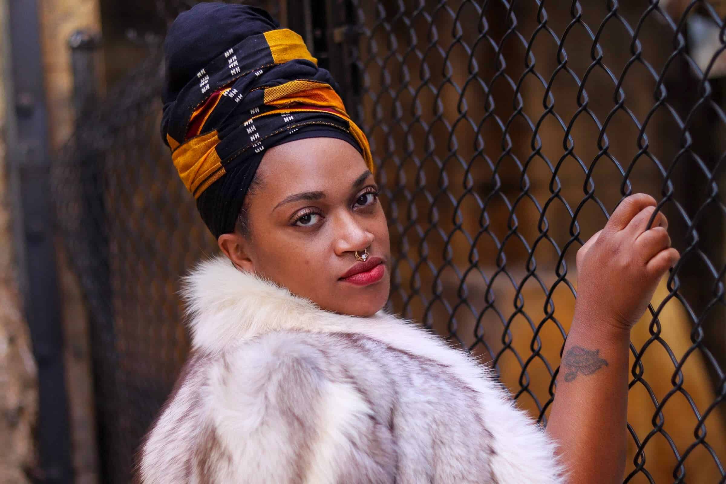Stylish woman with headwrap and fur coat standing by a chain-link fence.