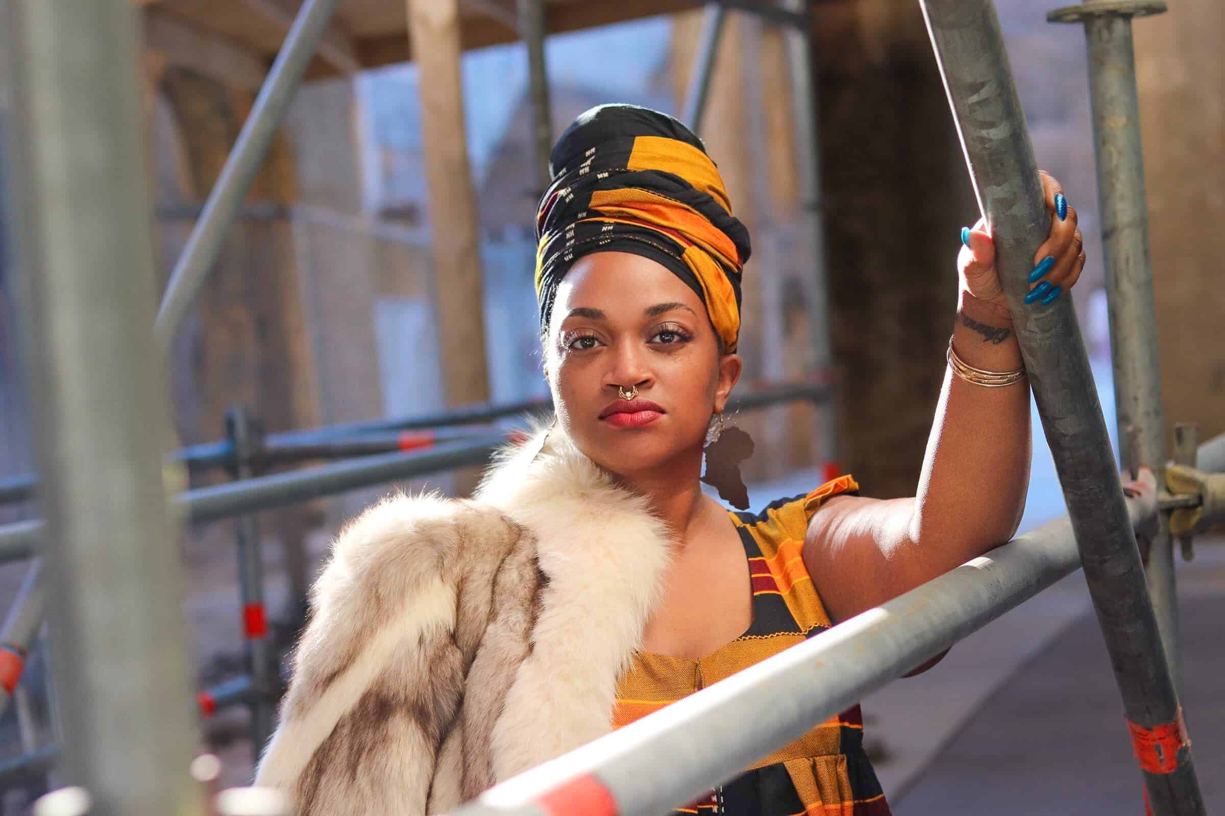 Confident woman with headwrap and fur coat standing on construction scaffolding.