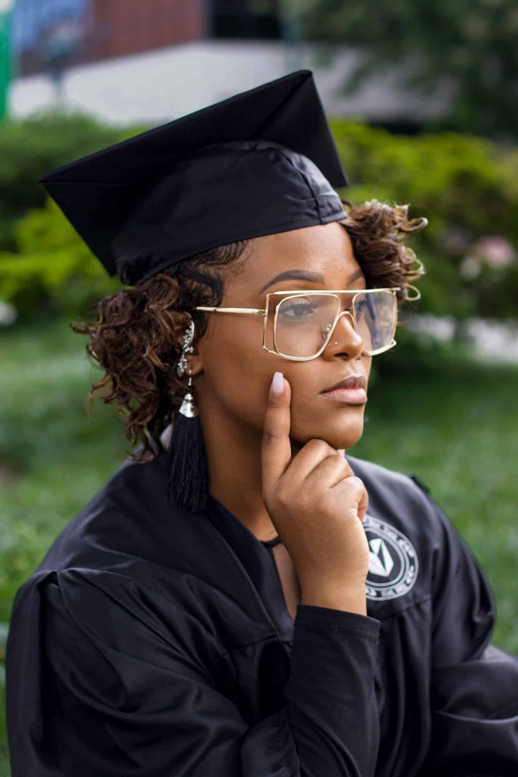 Graduate woman in cap and gown with glasses, outdoor background.