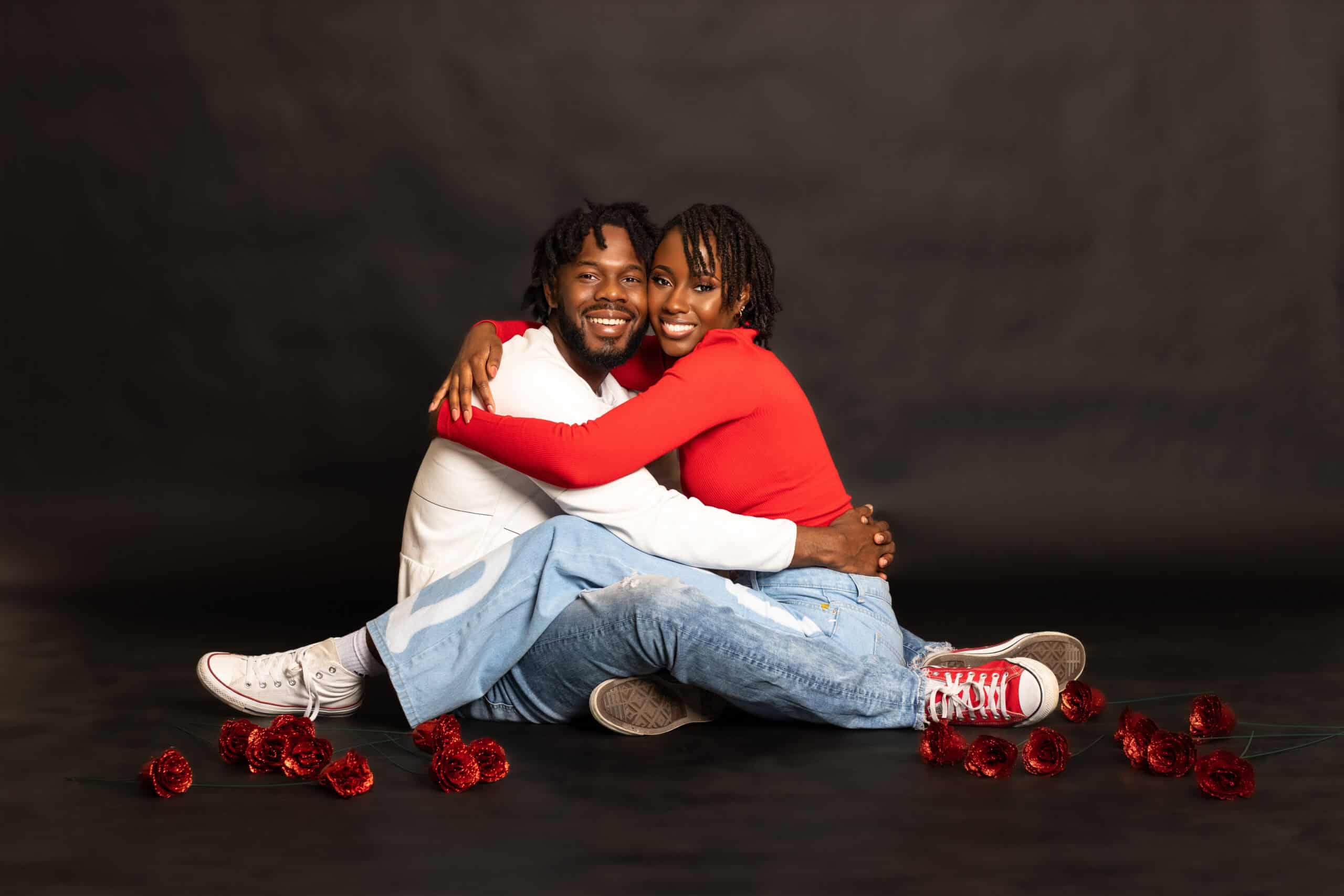 Joyful couple embracing on dark studio background with red roses around them.
