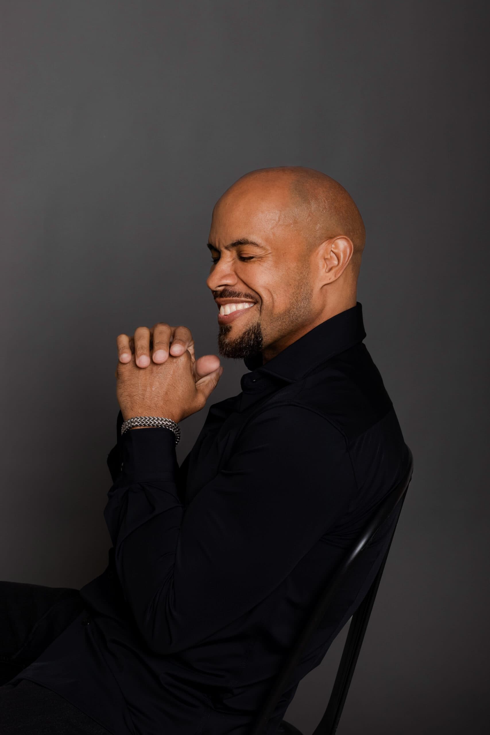 A smiling, confident man with a shaved head and beard, dressed in a black shirt, sitting against a d.