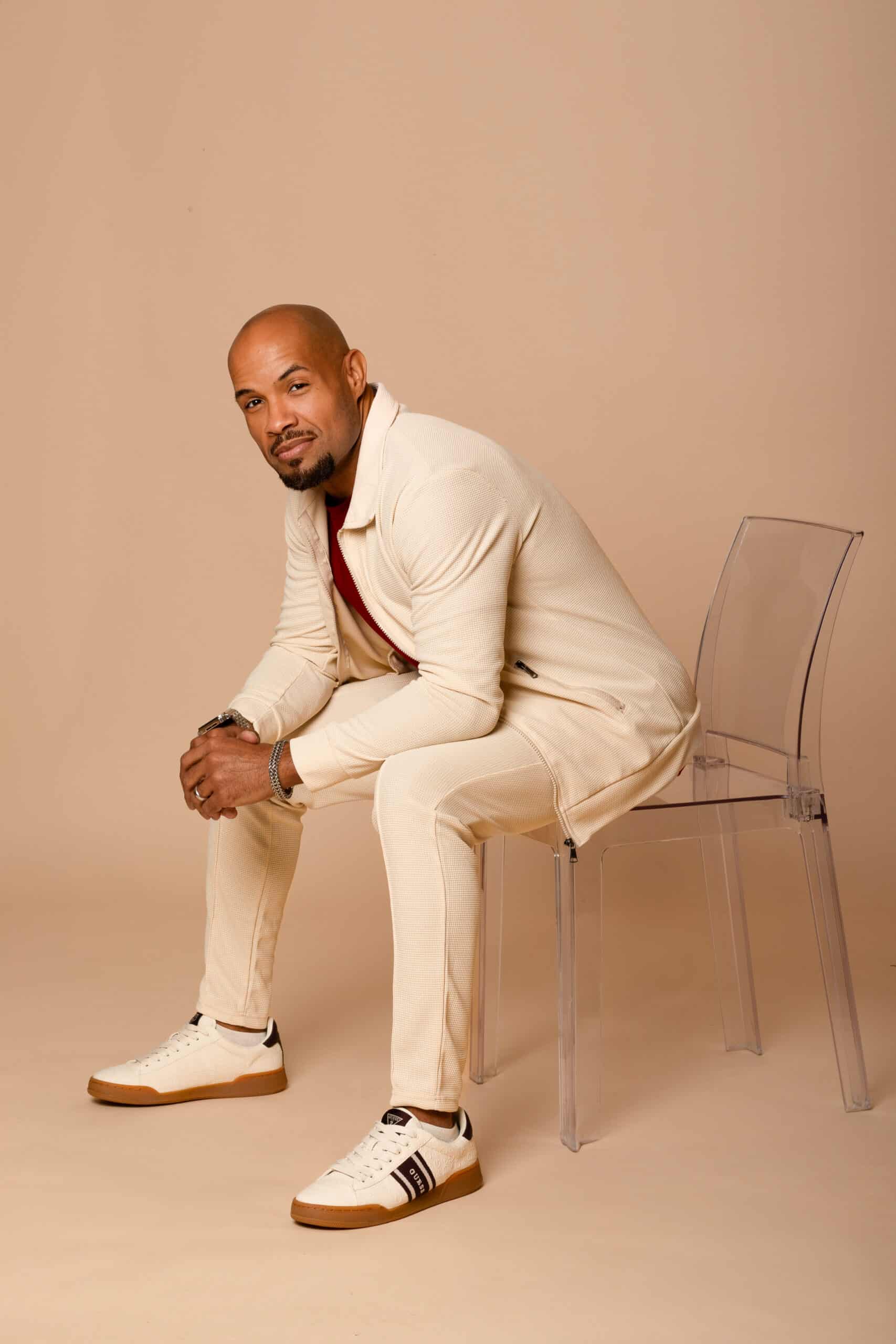 Man in beige suit sitting on transparent chair, casual pose, neutral background.