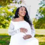 A pregnant woman smiling outdoors in a white lace dress with a floral headband.