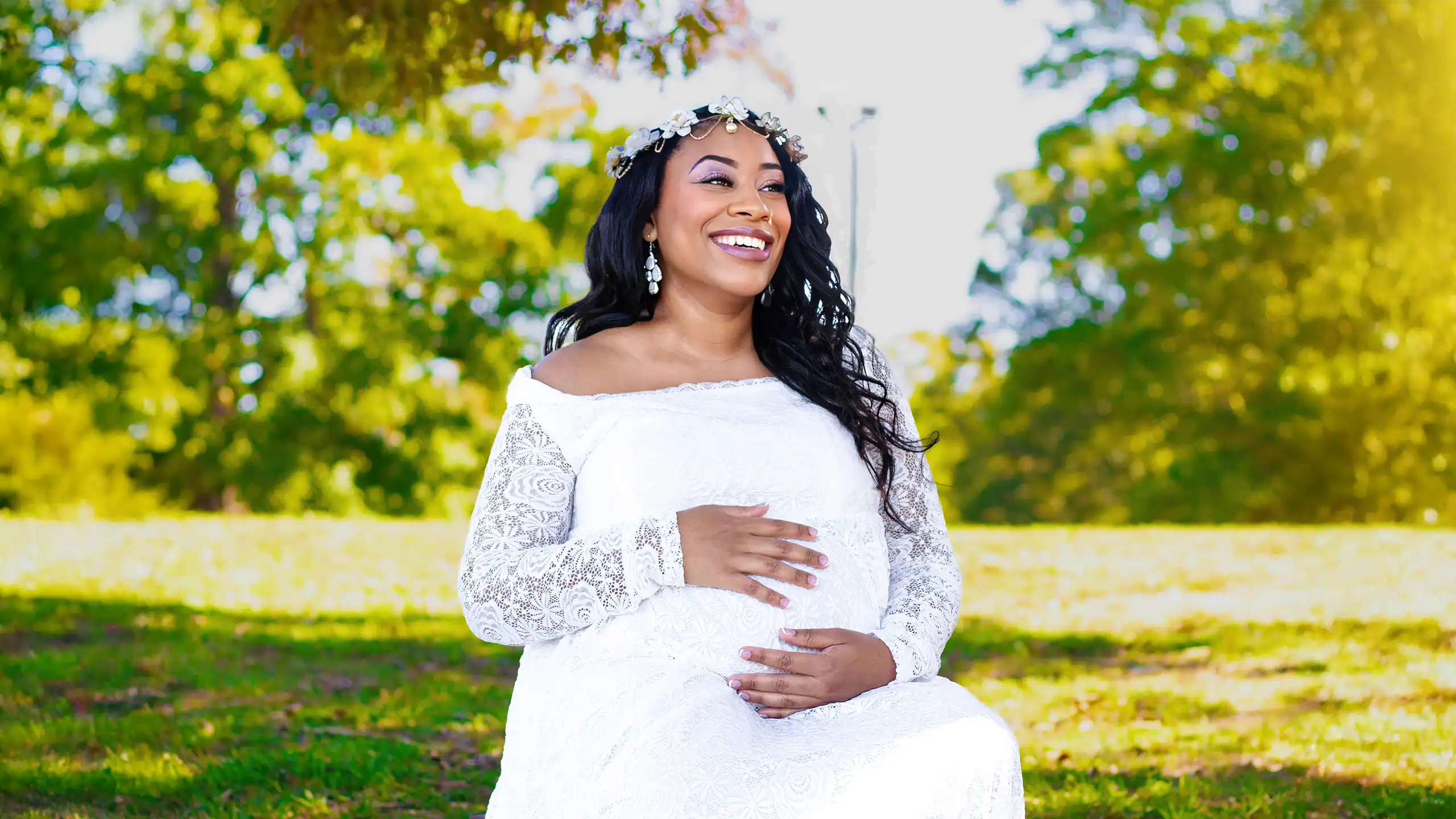 A pregnant woman smiling outdoors in a white lace dress with a floral headband.