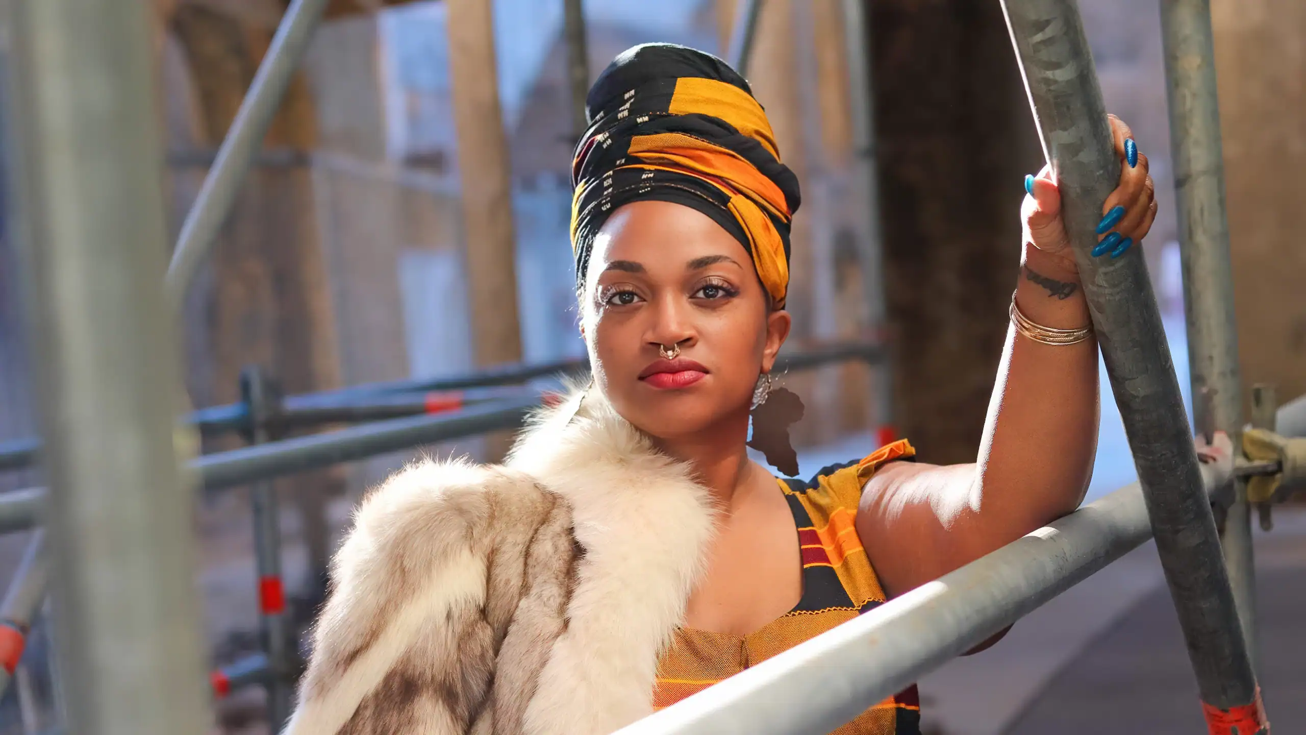 A woman with a colorful headwrap and fur stole posing on a construction site.