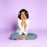 Bright smiling woman with curly hair sitting cross-legged on purple background, expressing happiness.