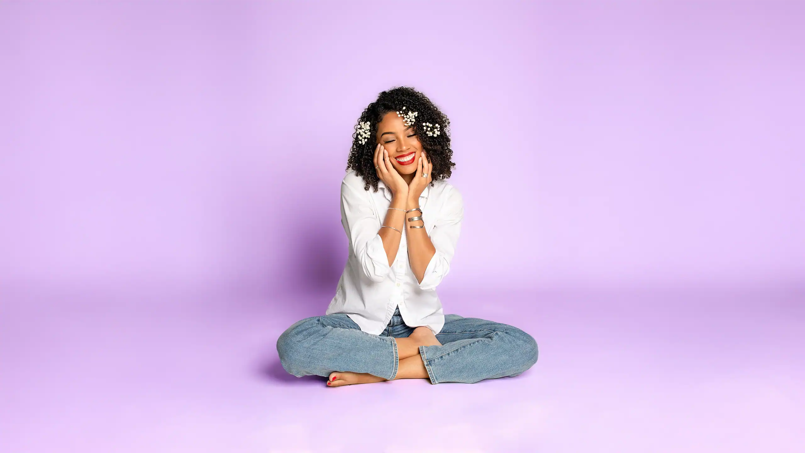Bright smiling woman with curly hair sitting cross-legged on purple background, expressing happiness.