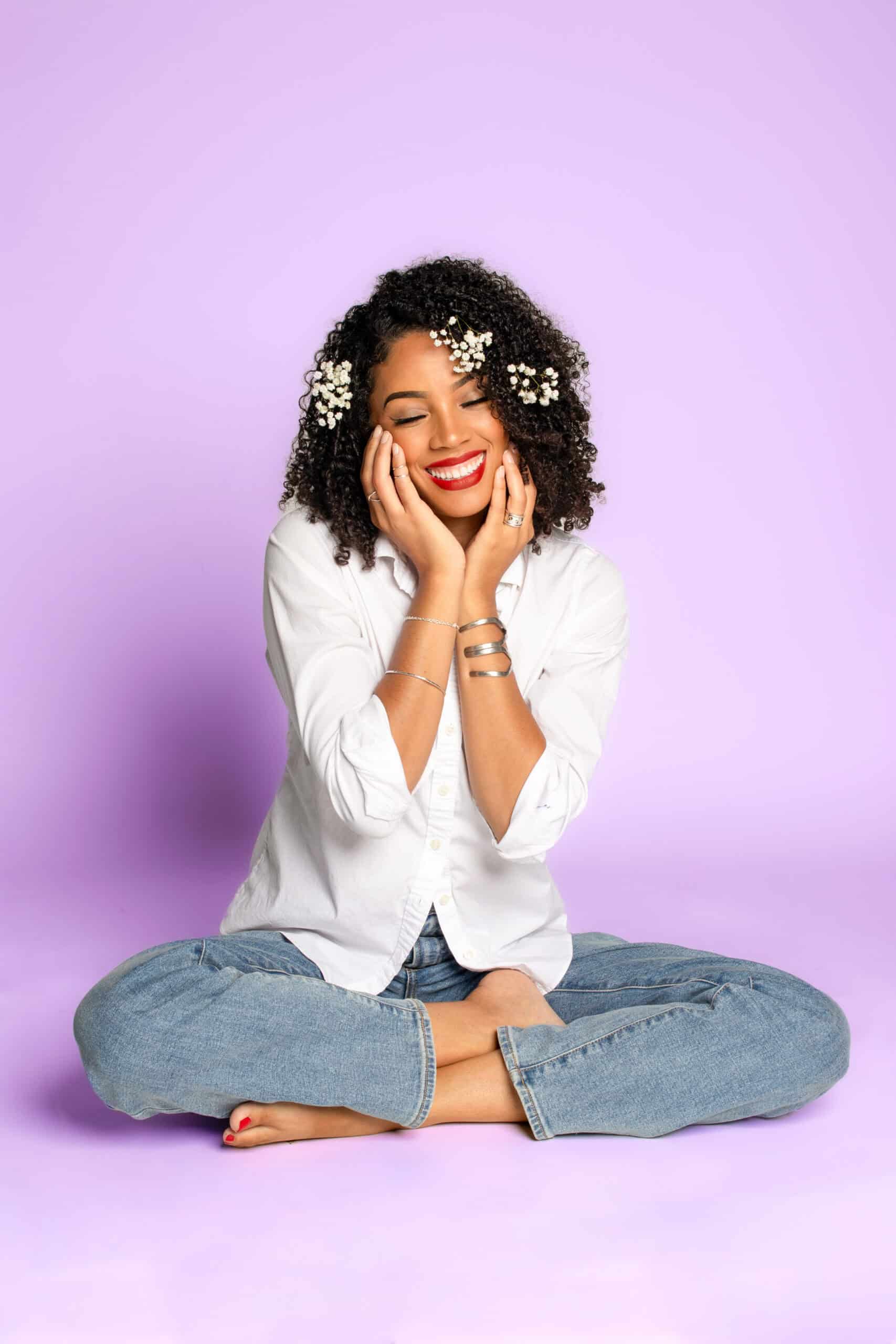 Smiling woman with flowers in curly hair, sitting cross-legged on purple background.